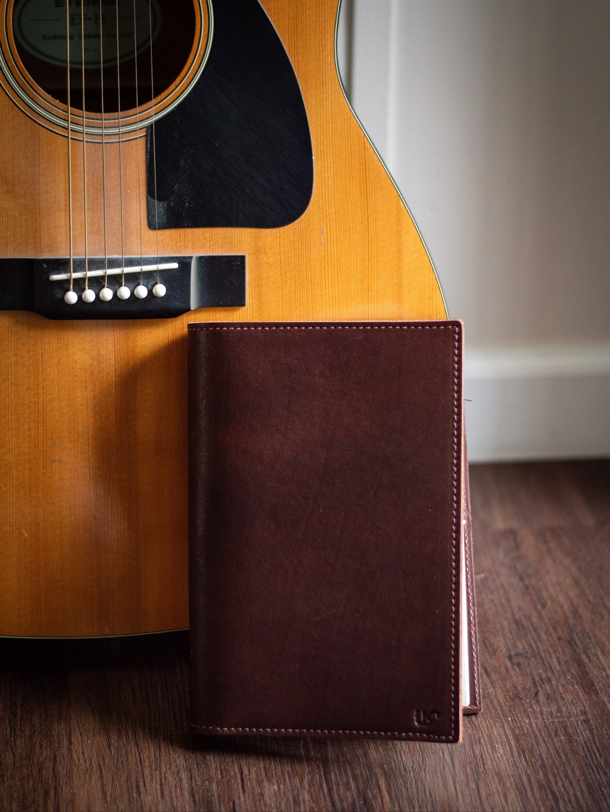 Acoustic guitar leans against a dark brown leather-bound notebook on a wooden floor.
