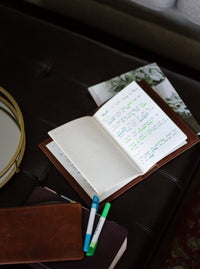 Open leather journal displays handwritten notes, resting on a dark leather surface with colorful highlighters and notebooks.
