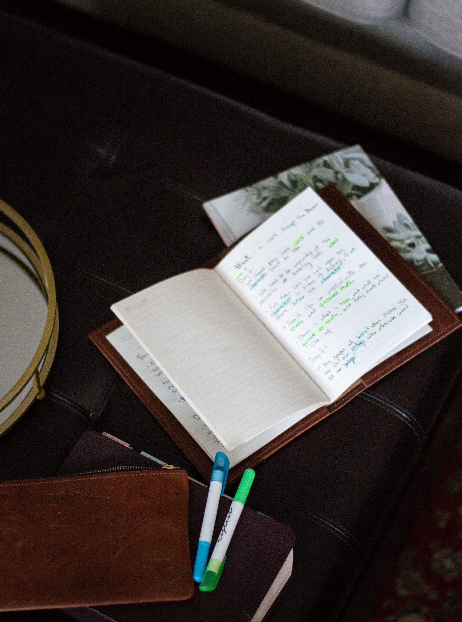 Open leather journal displays handwritten notes, resting on a dark leather surface with colorful highlighters and notebooks.
