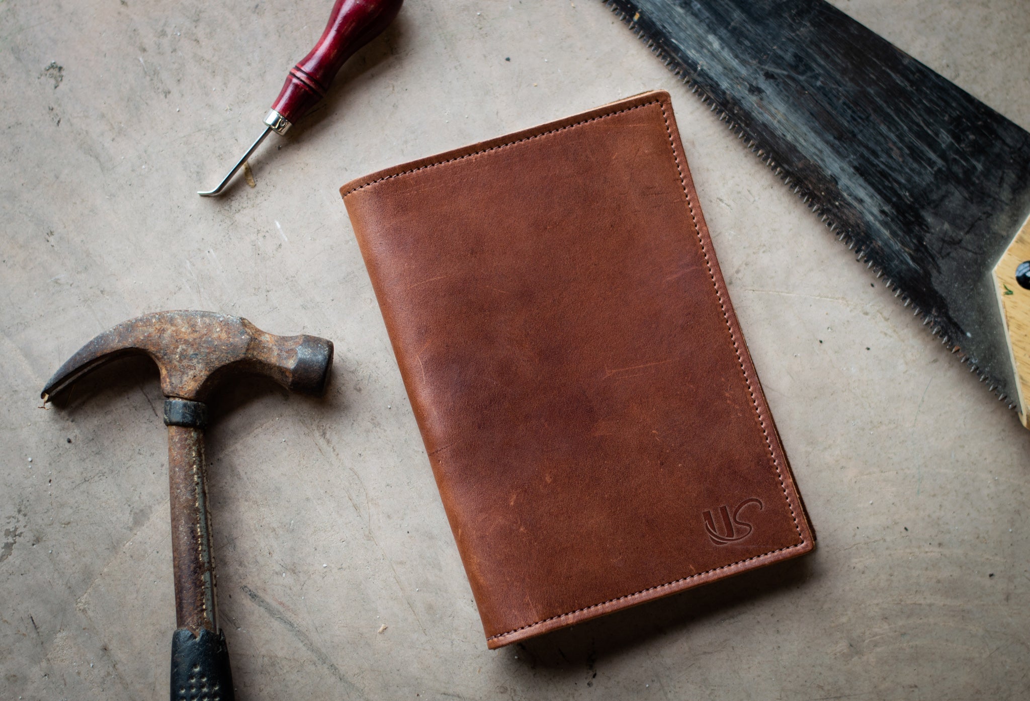 Craftspeople display handmade leather journal next to their tools: hammer, awl, and saw.
