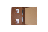 The opened brown leather journal displays two inserted photos and a pen, with blank paper ready for writing.