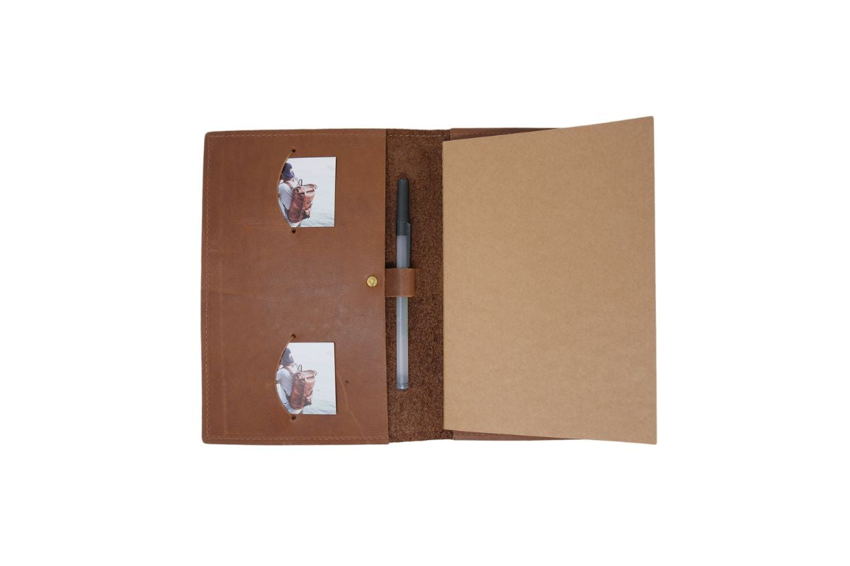 The opened brown leather journal displays two inserted photos and a pen, with blank paper ready for writing.
