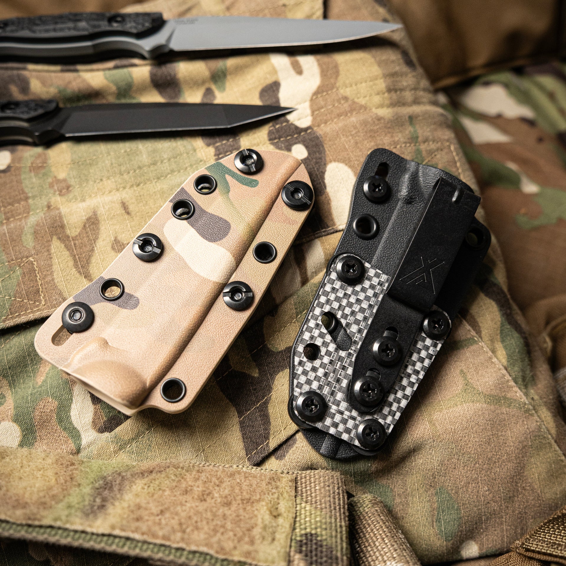 Two knives with different handles on a camouflage surface with two knife sheaths, one camouflage and one black.