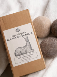 A package of hypoallergenic alpaca dryer balls, made in the USA, sits next to the felted wool balls on a white towel.
