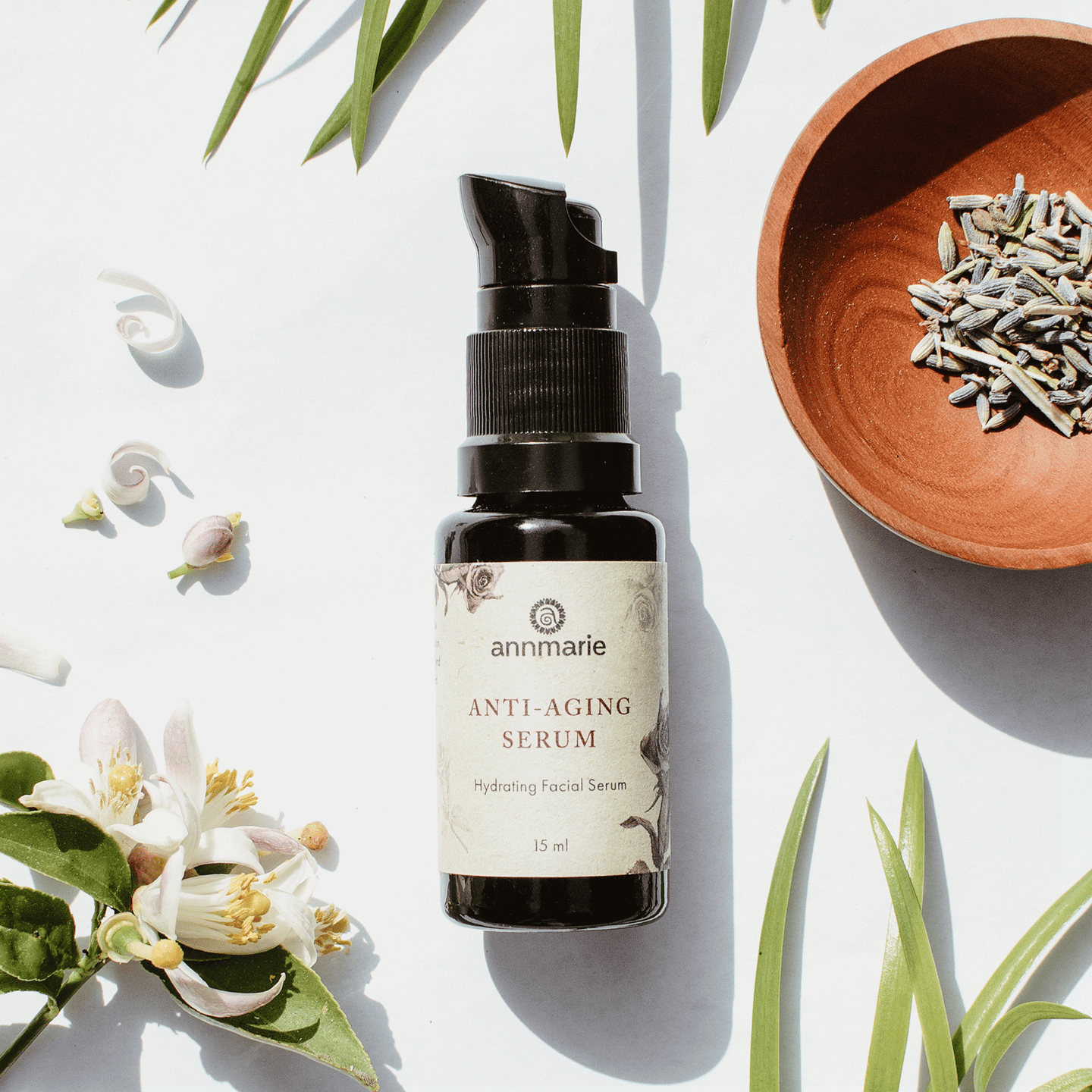 Annmarie Anti-Aging Serum sits on a white surface among sprigs, flowers, and lavender in a small wooden bowl.
