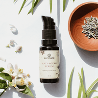 Annmarie Anti-Aging Serum sits on a white surface among sprigs, flowers, and lavender in a small wooden bowl.
