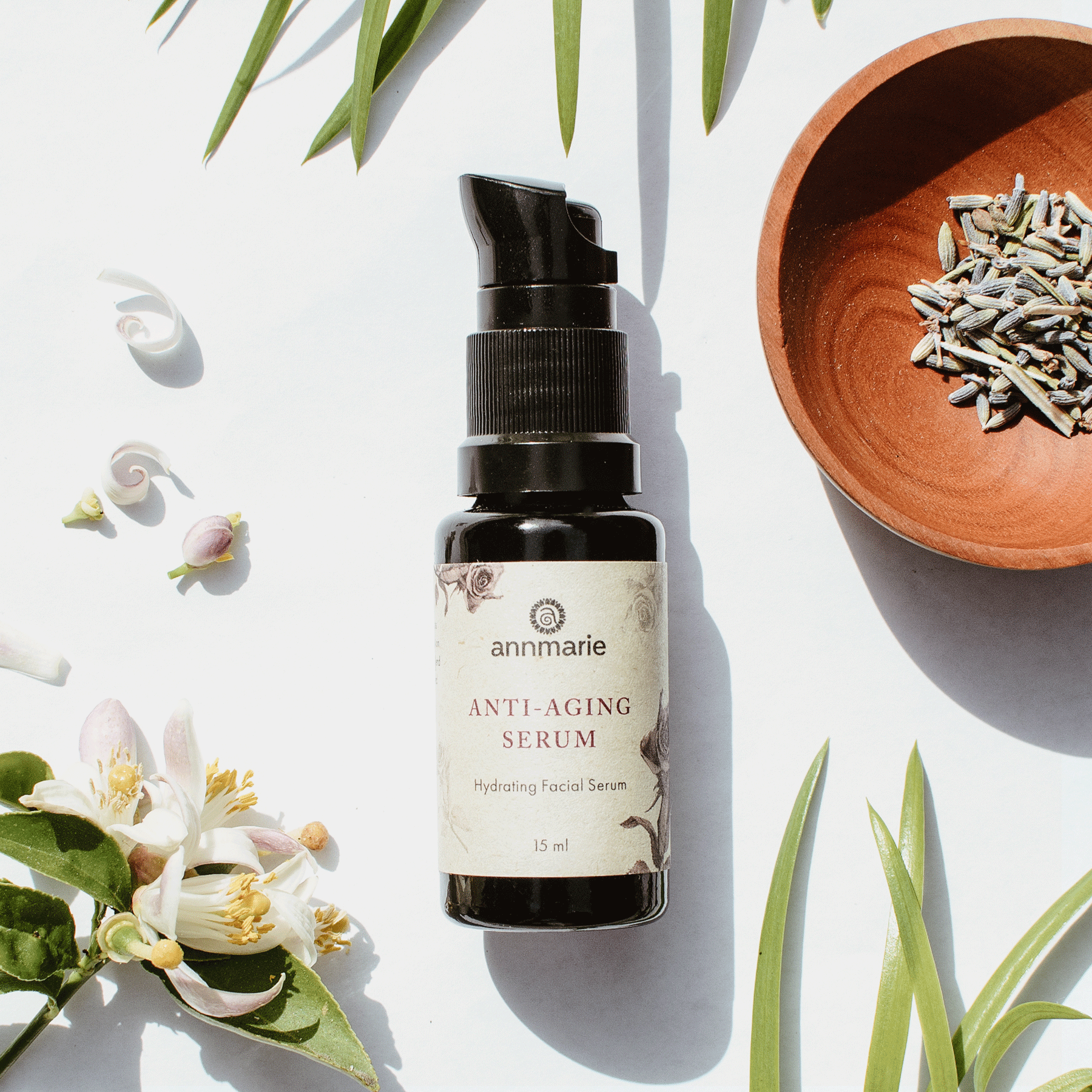 Annmarie Anti-Aging Serum sits on a white surface among sprigs, flowers, and lavender in a small wooden bowl.
