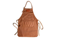 A light brown leather apron with pockets and adjustable neck strap lies flat against a white background.
