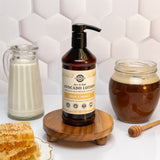 Rustic Strength Avocado Lotion, made with milk and honey, sits on a wooden stand next to milk, honey, and honeycomb.
