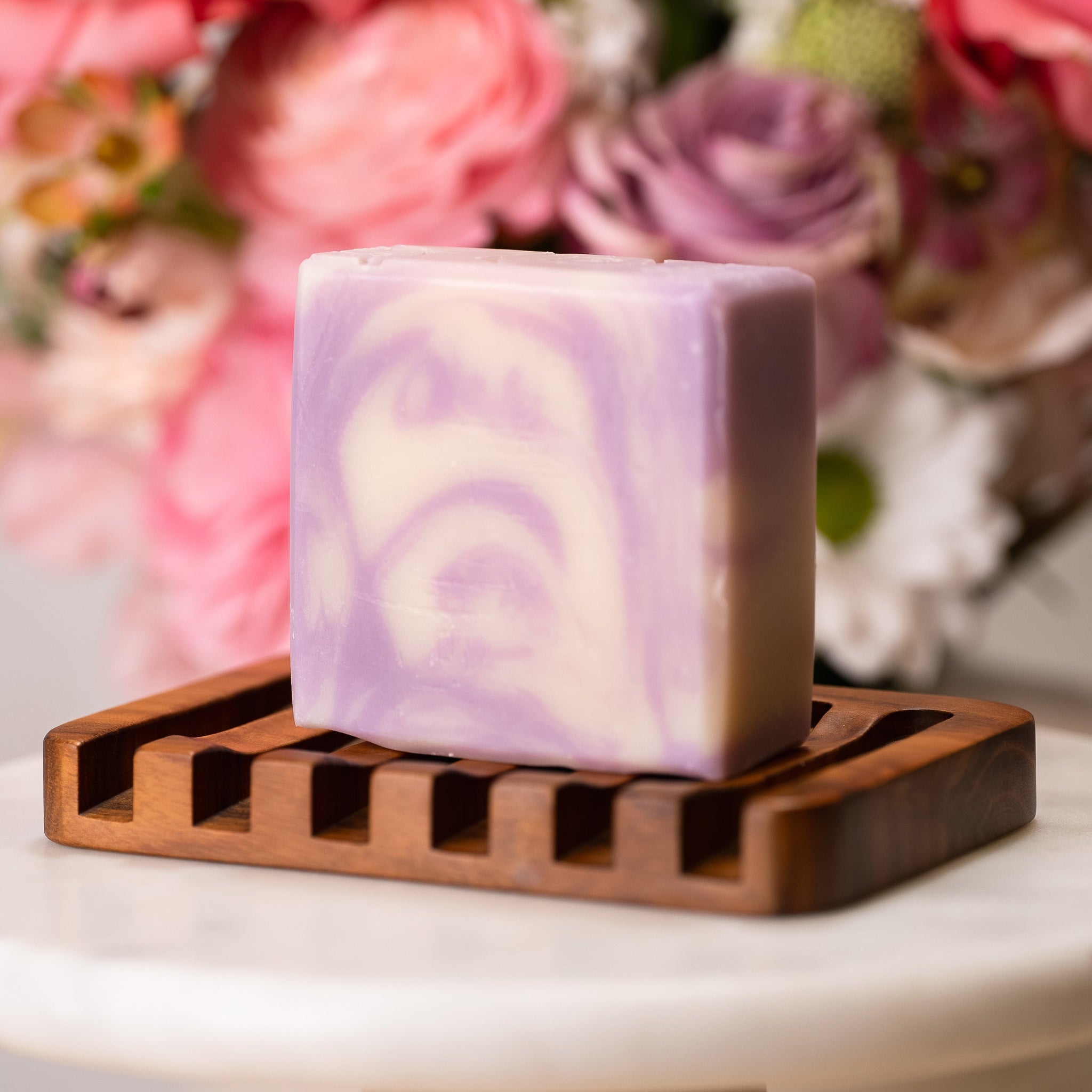 A lavender and white swirl soap bar rests on a wooden soap dish against a floral backdrop.
