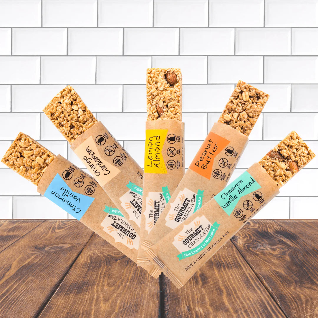 Six individually wrapped granola bars from The Gourmet Granola are fanned out on a dark wooden table in front of a white subway tile wall. The kraft paper wrappers show different flavor labels: Cinnamon Vanilla, Lemon Berry Blast, Peanut Butter, Cinnamon Vanilla Aromat, and Cinnamon Peanut Butter.