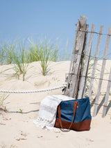 A stylish blue and brown beach bag sits on the sand next to a weathered fence and a striped towel on a sunny day.
