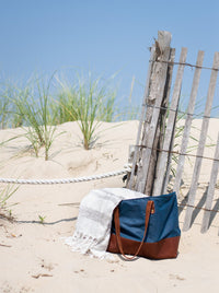A stylish blue and brown beach bag sits on the sand next to a weathered fence and a striped towel on a sunny day.
