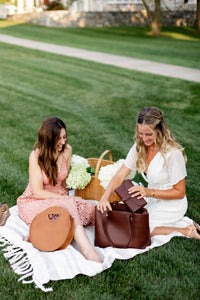 Two women enjoy a stylish picnic, with one taking a leather wallet from a brown leather tote bag while the other smiles.
