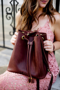 A woman in a pink lace dress holds a brown leather Urban Southern drawstring bucket bag.
