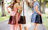 Three women model stylish bucket bags while walking on a tree-lined path, showcasing fashion and friendship.
