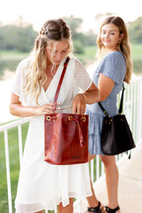 A woman models a stylish, oxblood leather bucket bag with drawstring closure while another woman carries a black leather bag in the background.
