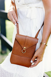 Woman displays a stylish brown leather crossbody bag with gold hardware, complementing her white dress and gold bracelet.

