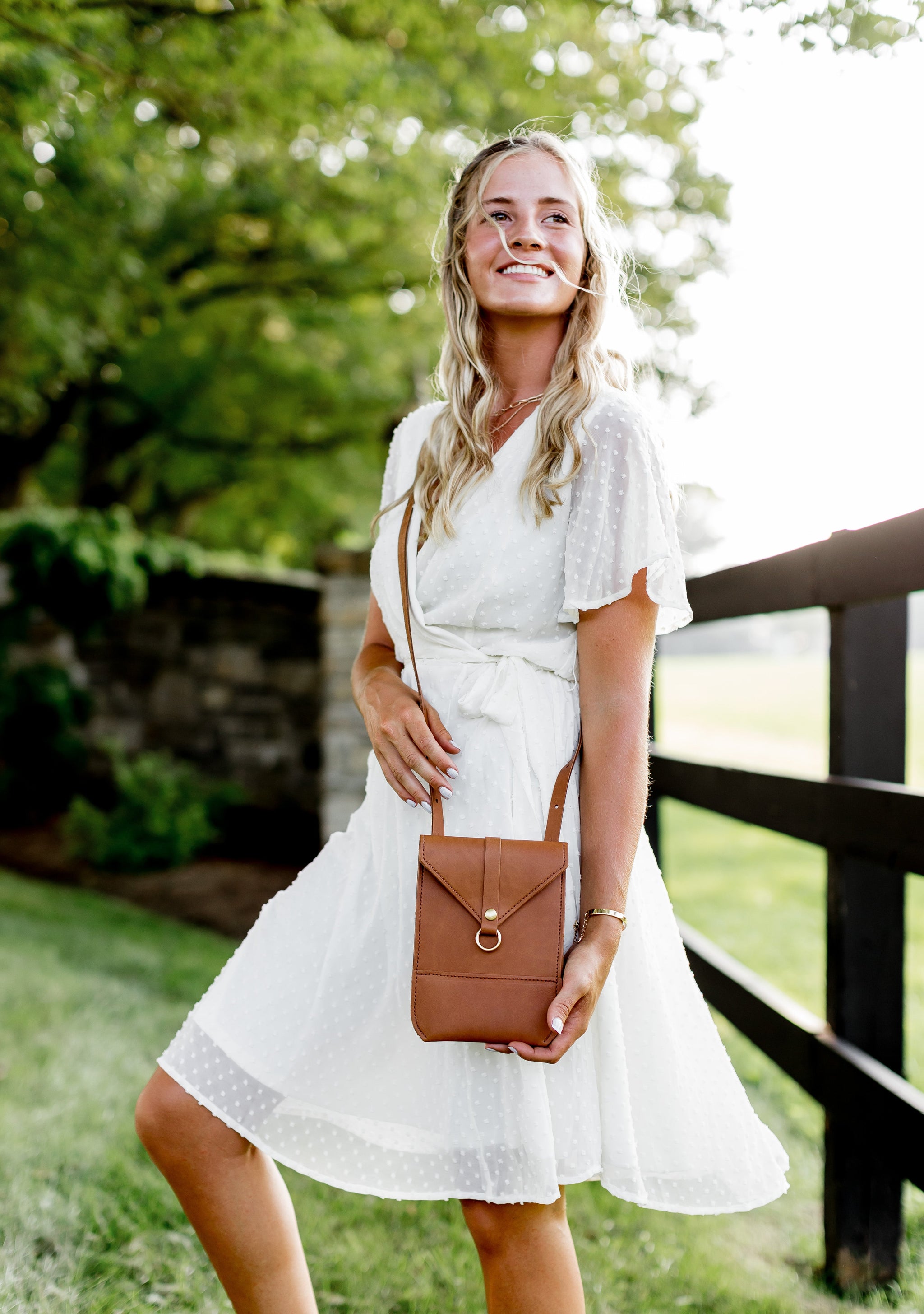 A blonde woman wearing a white dress and carrying a brown leather crossbody bag smiles outdoors.