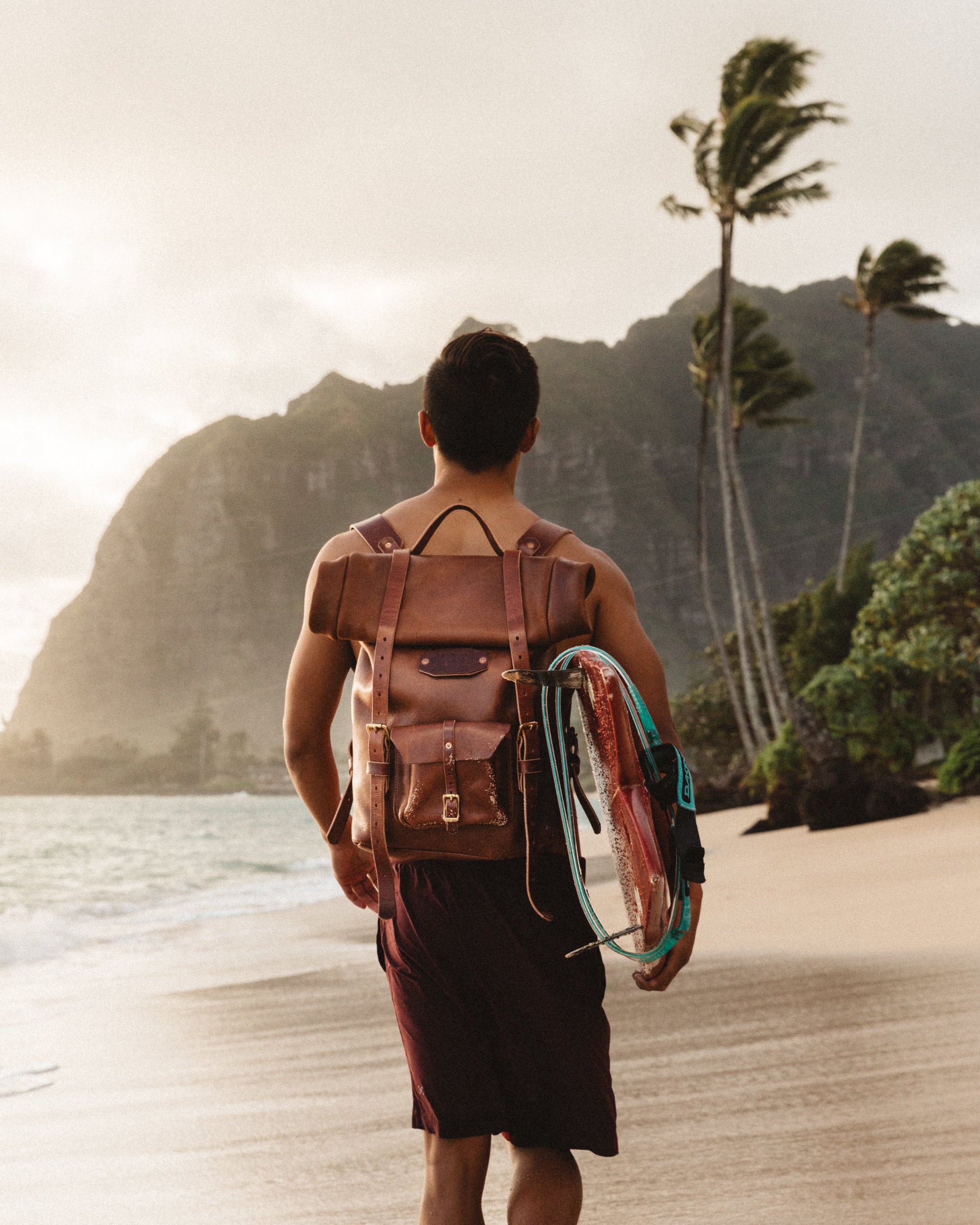 Man carries a vintage leather backpack and surfboard, walking towards the ocean on a tropical beach with palm trees and mountains.
