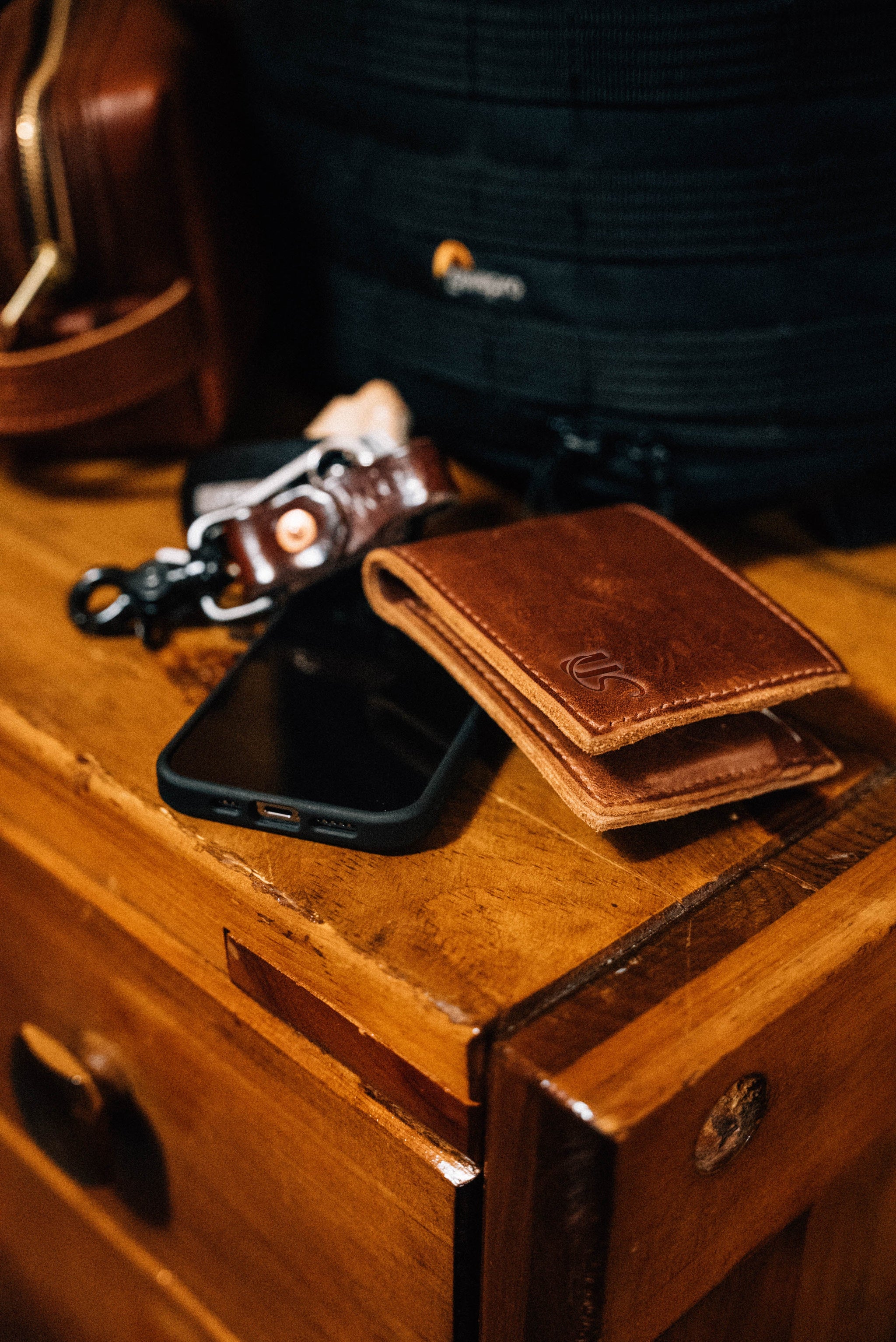 A brown leather wallet and smartphone rest on a wooden chest of drawers alongside a leather keychain and backpack.
