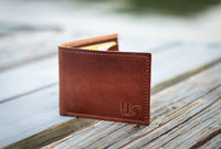 A brown leather wallet with "US" embossed sits on a weathered wooden surface, showcasing its craftsmanship.
