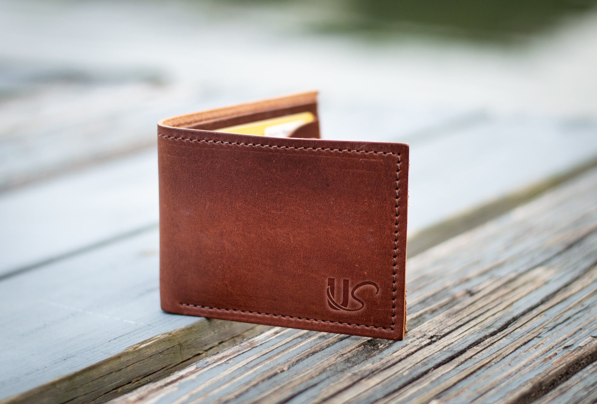 A brown leather wallet with "US" embossed sits on a weathered wooden surface, showcasing its craftsmanship.
