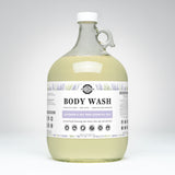View a one-gallon glass jug of Rustic Strength body wash featuring lavender and tea tree essential oils, promising luxurious lather and gentle cleansing for sensitive skin.
