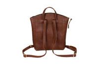 A brown leather backpack displays its adjustable straps, top handle, and zipped closure against a white background.
