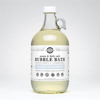 A clear glass jug displays Rustic Strength mama & baby safe unscented bubble bath, highlighting its vitamin B and cucumber extract ingredients.
