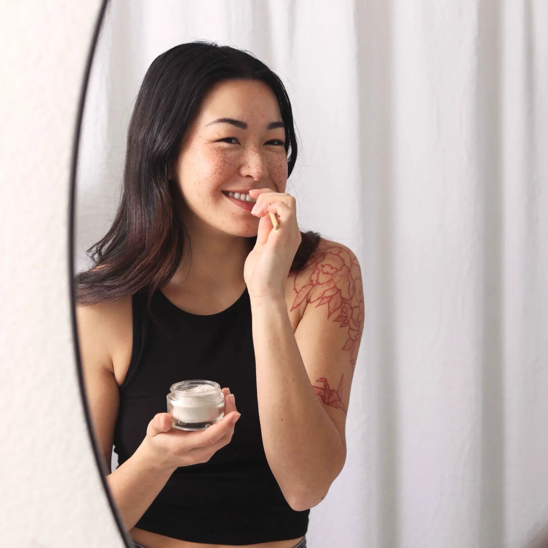 A woman with a floral tattoo smiles as she applies natural teeth whitening powder with a wooden applicator.
