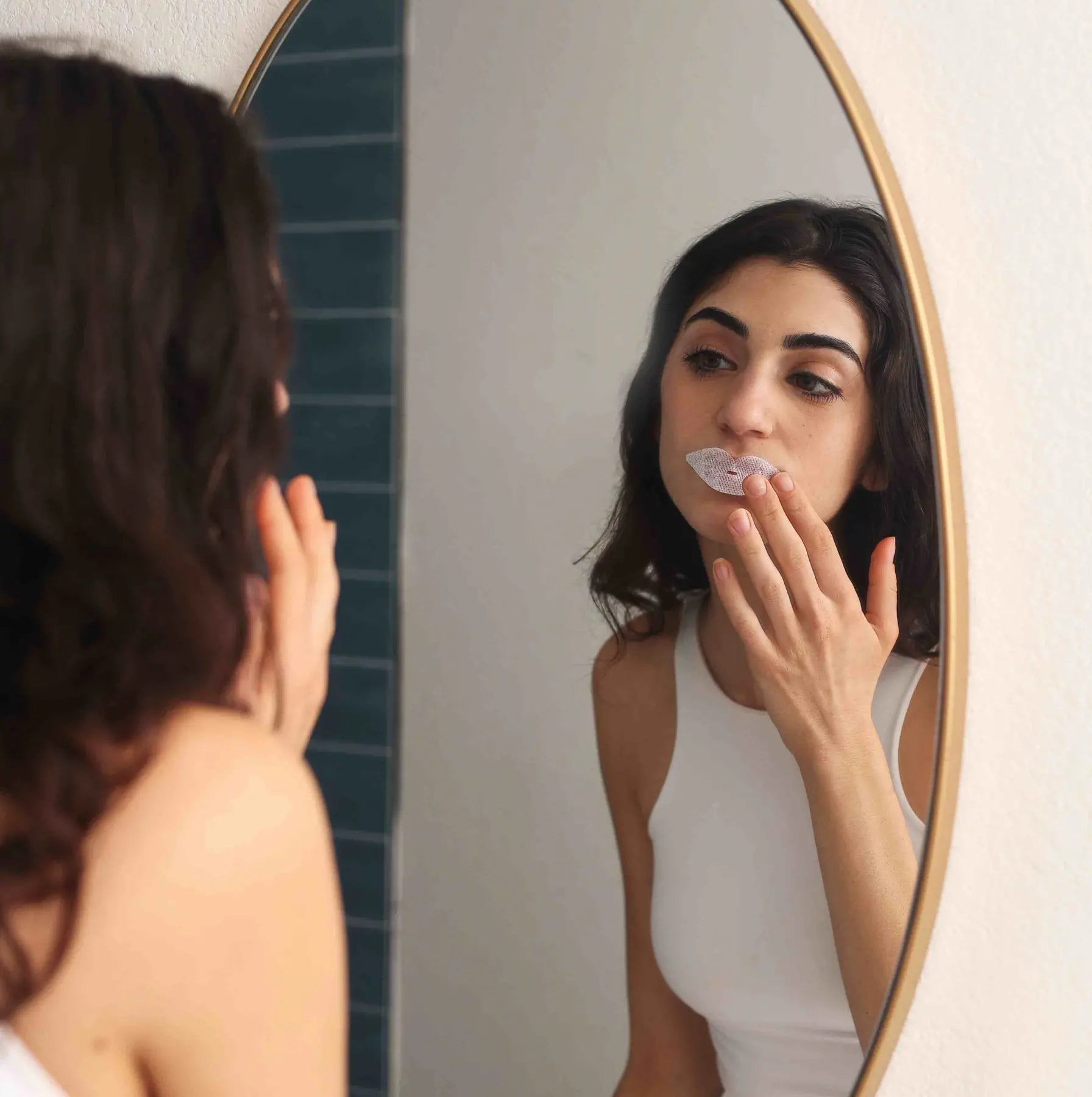 A woman applies a lip mask while looking in a gold-framed bathroom mirror.
