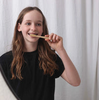 Teenage girl with braces brushes her teeth using a sustainable bamboo toothbrush in front of a mirror.

