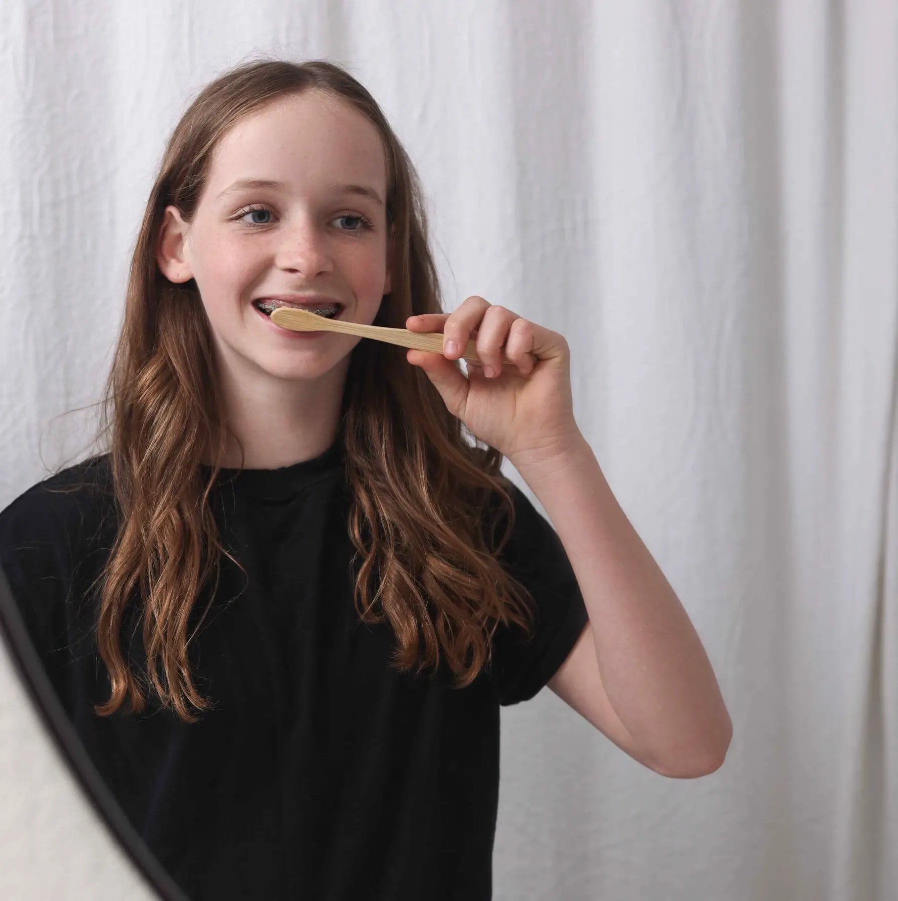 Teenage girl with braces brushes her teeth using a sustainable bamboo toothbrush in front of a mirror.
