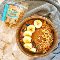 Bowl of colorful cereal with bananas and a package of 'The Gourmet Crumbles' on a marble surface.