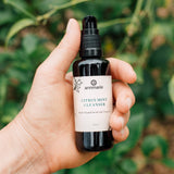 Hand holds a bottle of Annmarie Citrus Mint Cleanser, an herb-infused facial gel cleanser, against a blurred green foliage background.

