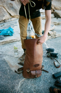 A rock climber packs quickdraws and gear into a leather climbing pack on a granite boulder.
