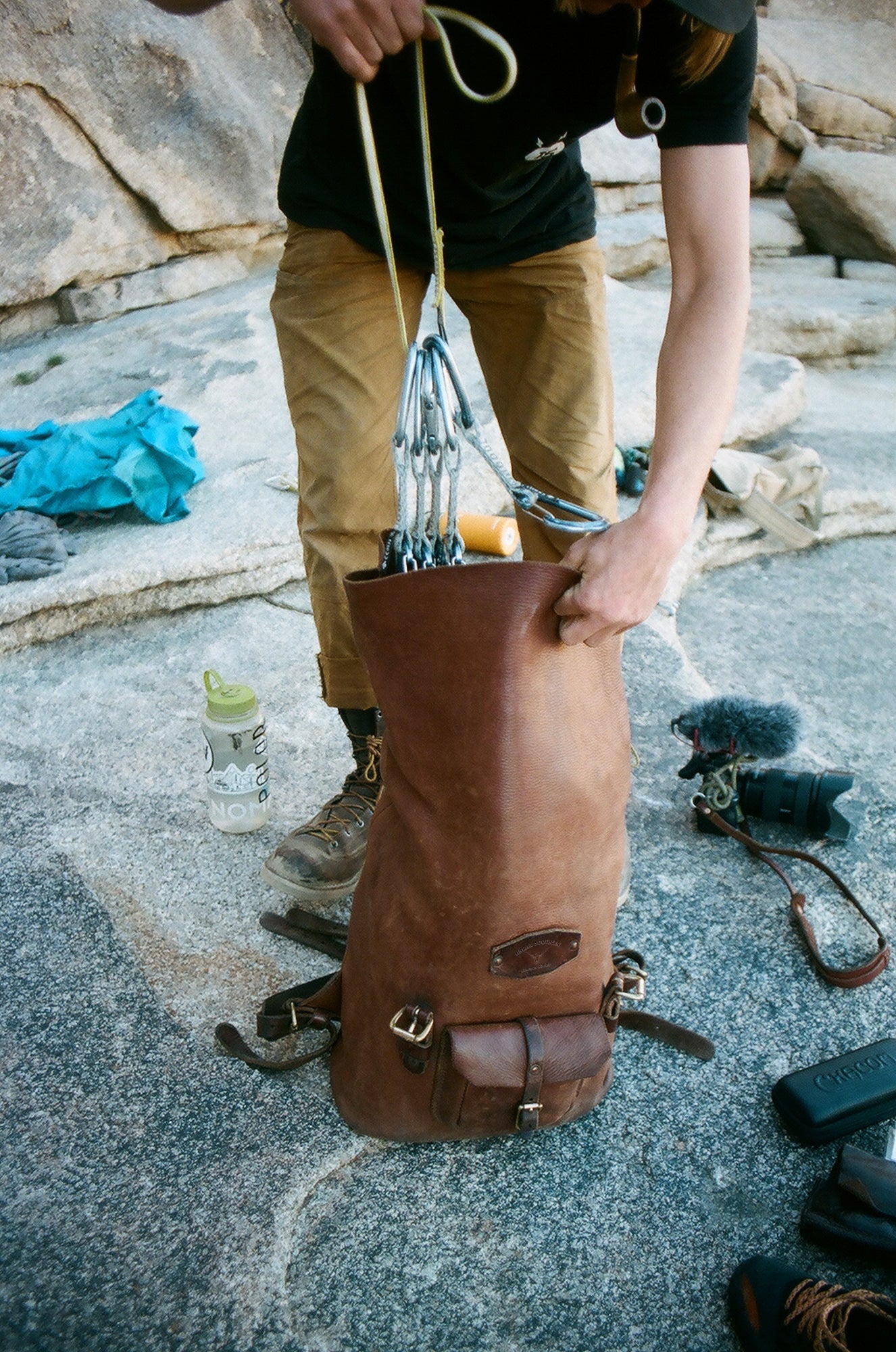 A rock climber packs quickdraws and gear into a leather climbing pack on a granite boulder.
