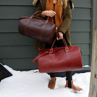Person holding two leather bags, one brown and one red, against a snowy background.