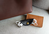 A black leather Urban Southern cord organizer keeps white charging cables tidy next to a brown leather wallet and bag.
