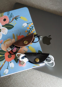 Brown and black leather cable organizers keep charging cords tidy on a floral notebook beside a Macbook.
