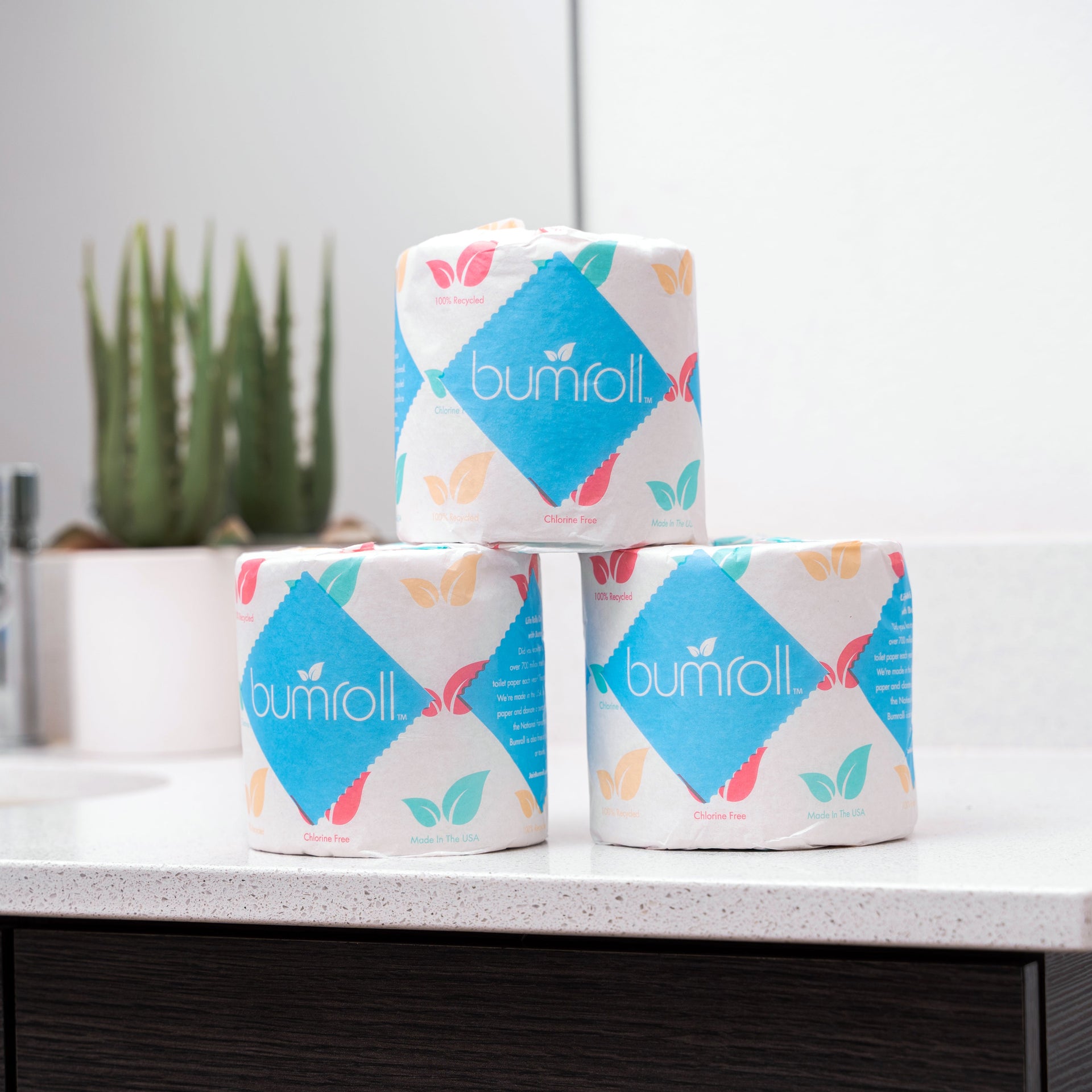 Three rolls of eco-friendly Bumroll toilet paper, boasting chlorine-free and recycled materials, stack on a modern bathroom counter.