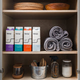 Organized shelves display Bloop laundry soap, rolled towels, cleaning supplies, and wooden bowls in a light wood cabinet.
