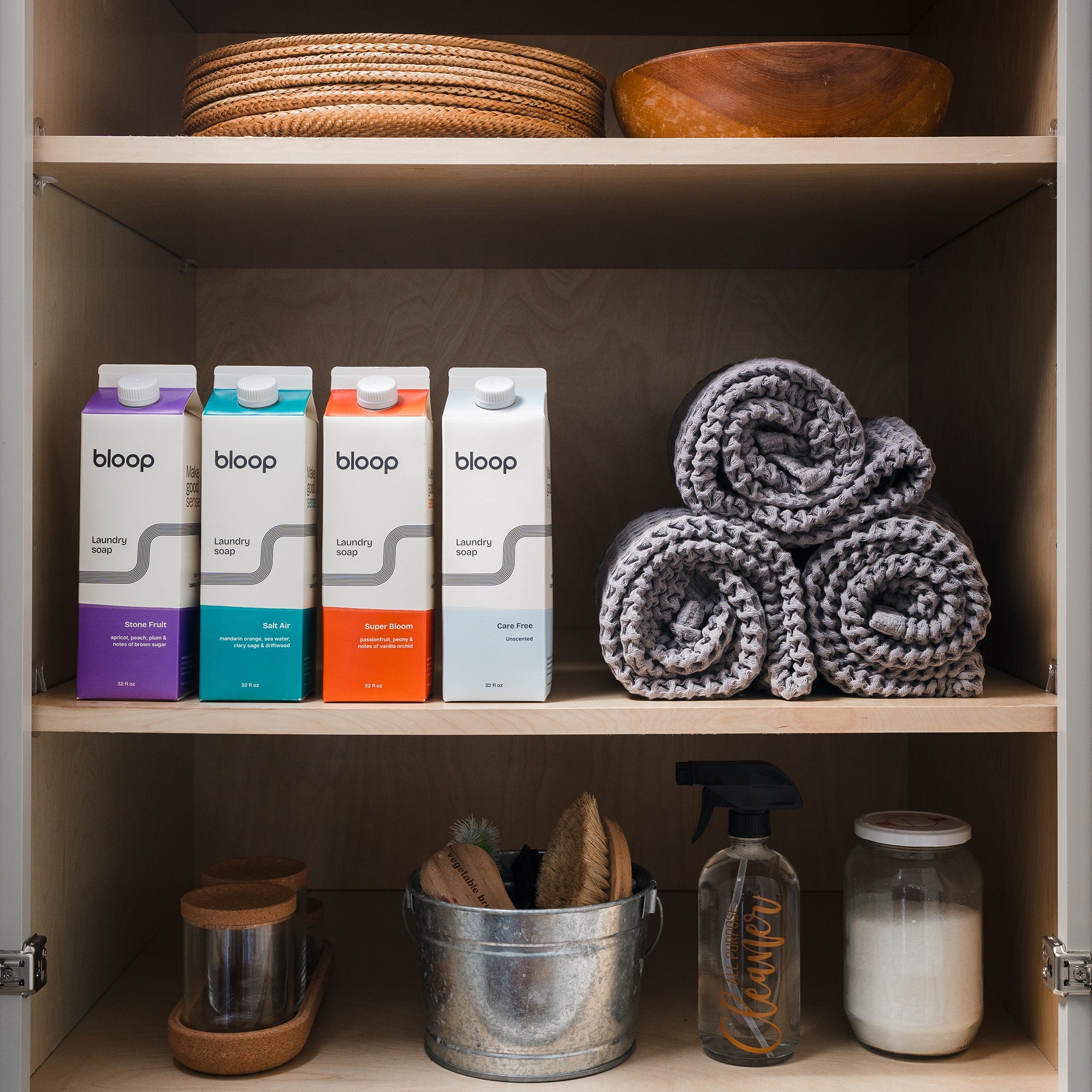 Organized shelves display Bloop laundry soap, rolled towels, cleaning supplies, and wooden bowls in a light wood cabinet.
