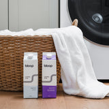 Bloop laundry soap cartons sit near a woven basket overflowing with white towels, showcasing eco-friendly laundry solutions.
