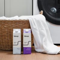 Bloop laundry soap cartons sit near a woven basket overflowing with white towels, showcasing eco-friendly laundry solutions.
