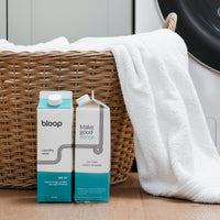 Two cartons of eco-friendly Bloop laundry soap sit beside a wicker basket overflowing with white towels in a laundry room.