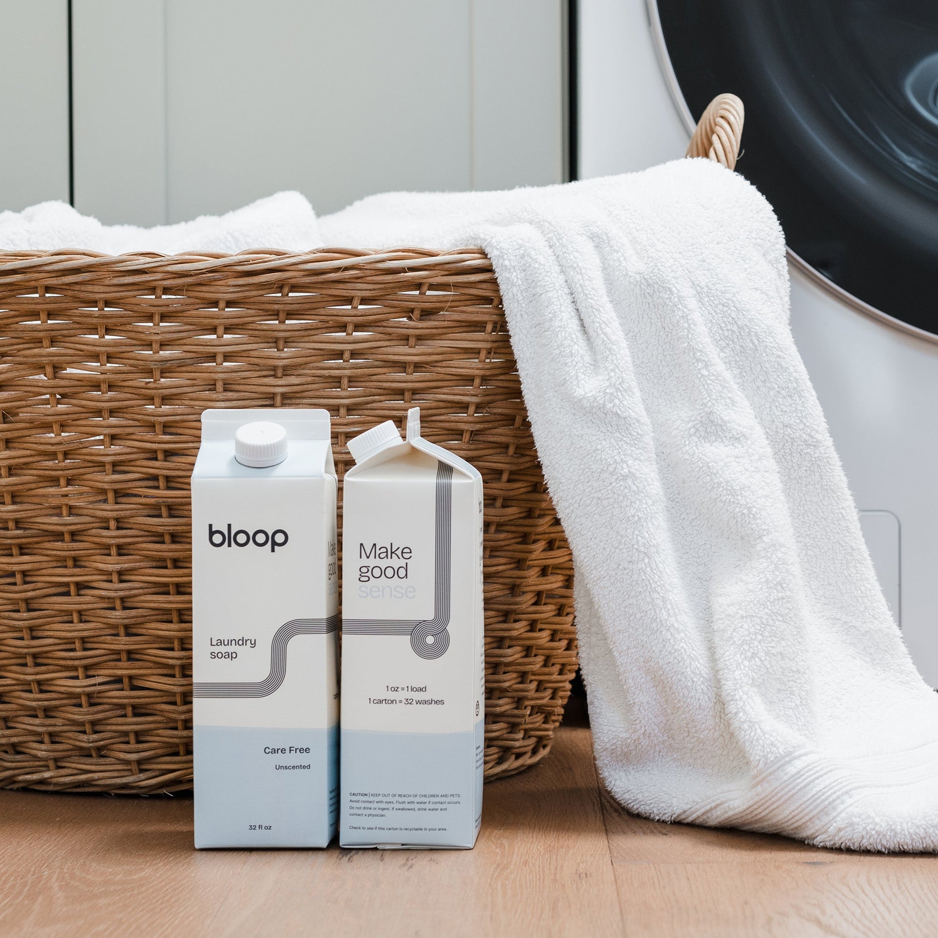 Two cartons of eco-friendly "bloop" laundry soap sit near a wicker basket overflowing with fresh, white towels in a modern laundry room.

