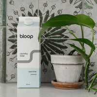 Showcases Bloop unscented laundry soap next to a potted plant against a patterned tile backdrop.

