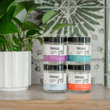 Blooop laundry soap jars, including scents like Stone Fruit and Salt Air, stack on a wooden shelf alongside a potted plant.
